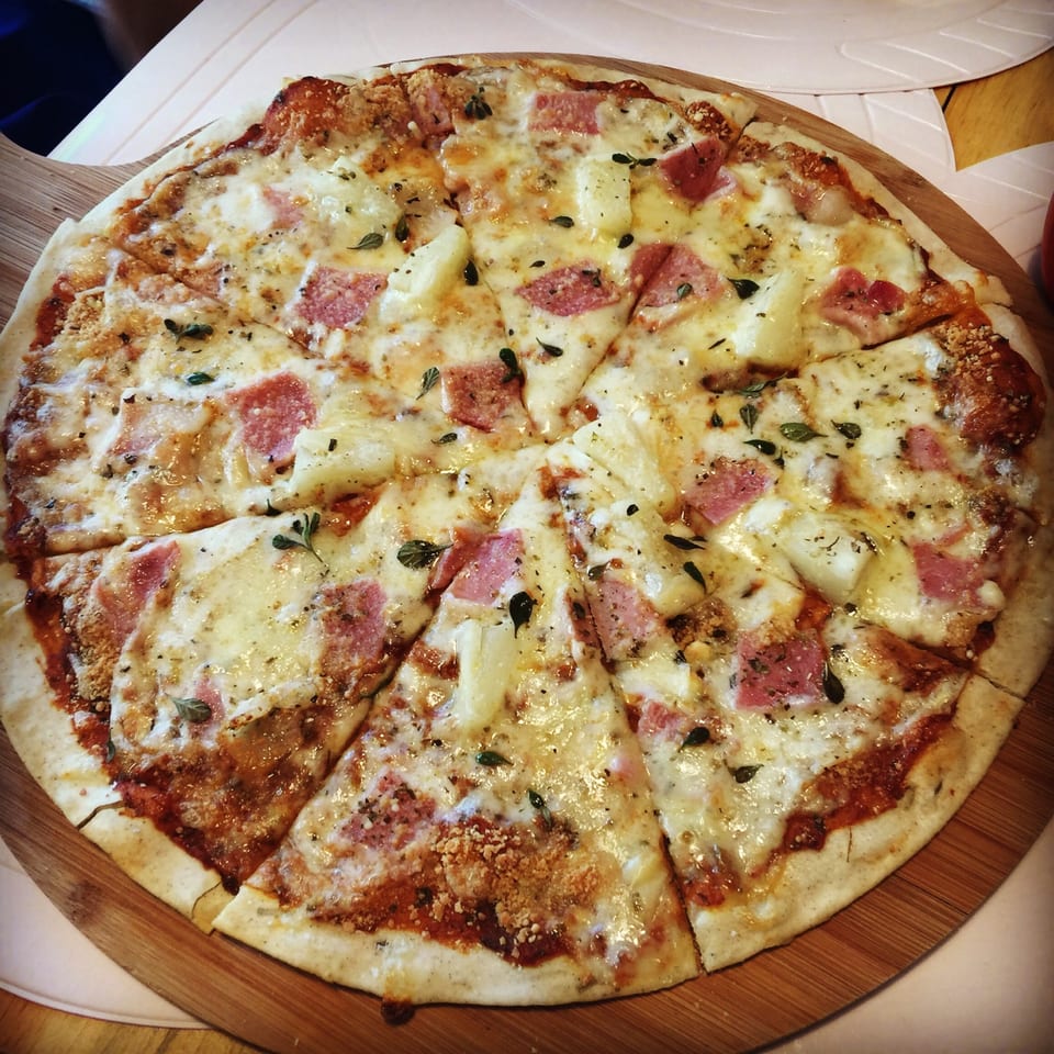 A freshly baked Pizza 24 pizza, topped with ham, pineapple, cheese, and herbs, on a wooden board.